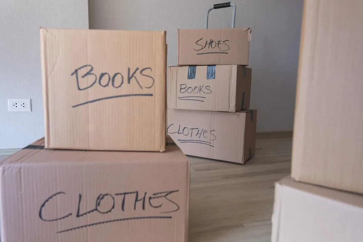 Stacked moving boxes labeled for books, clothes, and shoes inside a new home, on light wood flooring.